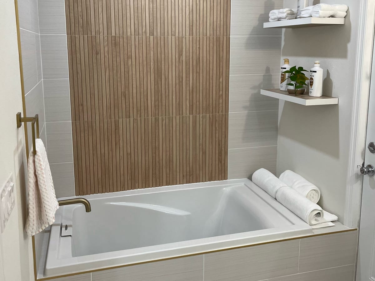 Soaking tub beneath a wood-slat feature wall with brushed-gold fixtures and floating shelves