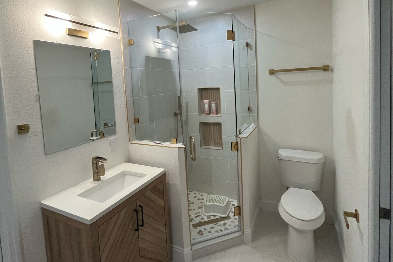 Corner shower with frameless glass, brass hardware and a terrazzo floor, beside a chevron wood vanity